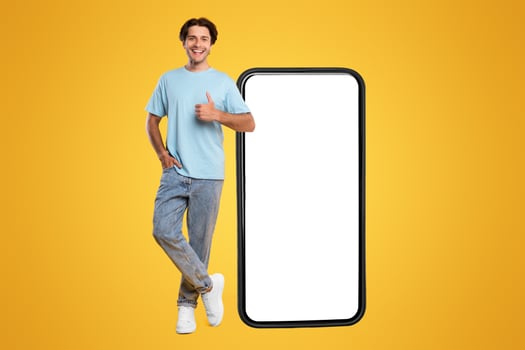 Happy Man Standing Near Big Smartphone With Empty White Display Gesturing Thumb Up Sign, Cheerful Casual Male Recommending New App Or Website, Standing On Yellow Background, Mock Up, Full Body Length photo
