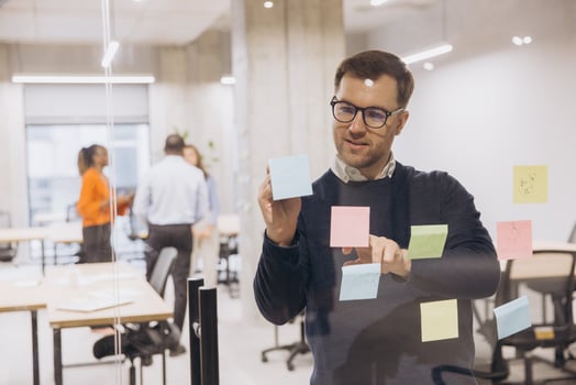 Businessman using sticky notes on a glass wall in a modern office, collaborating with colleagues in the background to brainstorm ideas photo