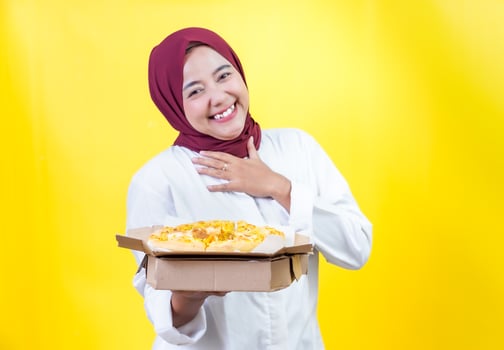 A joyful asian woman in a hijab presents a delicious pizza against a bright yellow background photo