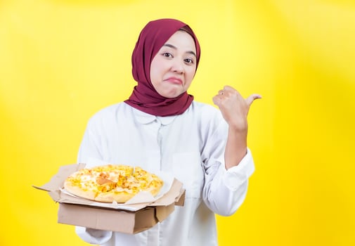 An asian woman in a hijab holds a pizza box and points back with her thumb against a yellow background. photo