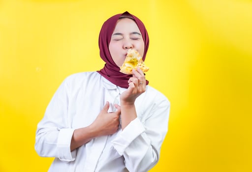 A joyful asian woman in a hijab savors a delicious slice of pizza against a bright yellow backdrop. photo