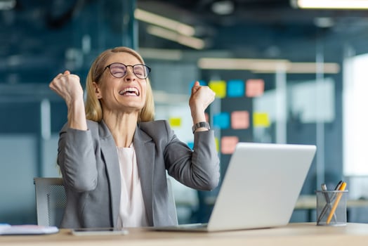 Joyful businesswoman in office celebrating achievement with clenched fists. Displays excitement and success. Sitting at desk with laptop, this professional embodies career accomplishment photo
