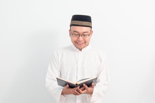Smiling Asian Muslim man wearing white clothes reading quran during Ramadan Eid Al Fitr and eid adha days celebration isolated white background. Islamic celebration concept. photo