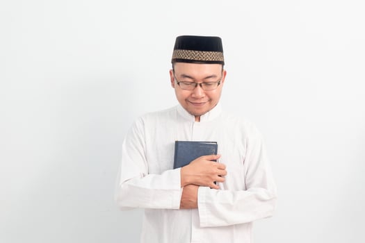 Cheerful Asian Muslim man wearing white clothes holding quran during Ramadan Eid Al Fitr and eid adha days celebration isolated white background. Islamic celebration concept. photo