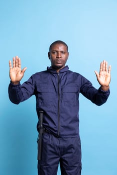 Security agent raising his hands up as a stop symbol in studio, restricting the access with a forbidden sign against blue background. Serious bodyguard in uniform does a limitation gesture. photo