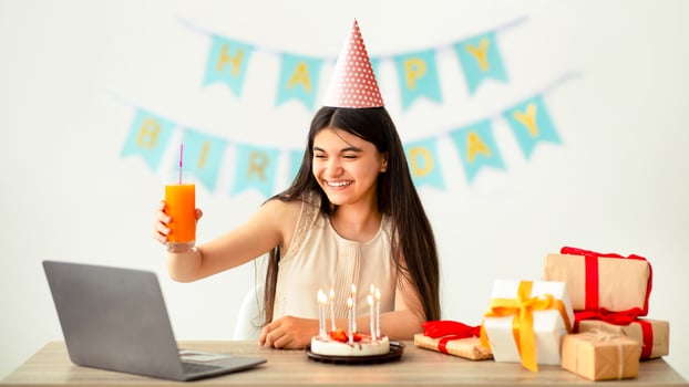 Cool Indian teen girl having online birthday party from home during coronavirus lockdown, wearing festive hat, holding glass of juice, saluting at laptop screen, conferencing with her friends photo