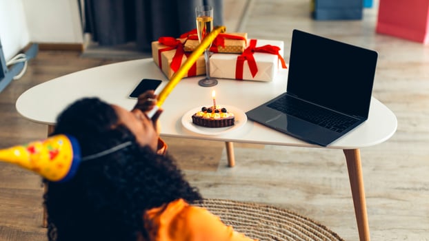 Online Birthday Concept. African American Lady At Laptop Blowing Party Horn Celebrating B-Day Holiday Making Call At Home, Wearing Festive Hat. Selective Focus On Computer, Mockup, Rear View photo