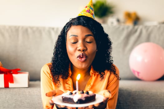 African American Woman Making Birthday Wish And Blowing Candle On Cake Wearing Festive Hat Sitting With Eyes Closed At Home. B-Day Holiday Celebration Concept. Front View photo