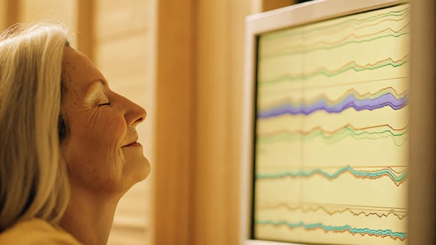 A side view of a senior participant eyes closed and a slight smile as colorful graphs appear on a computer monitor indicating positive brainwave activity and focus during training.. photo