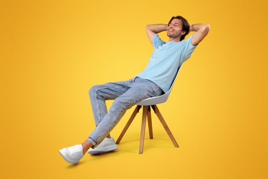 Vertical Portrait Of Happy Casual Millennial Man Relaxing Sitting In Chair Holding Hands Behind Head Leaning Back, Posing With Eyes Closed On Yellow Orange Studio Wall Background, Enjoying Silence photo