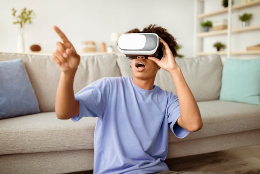 Contemporary stay home pastimes. Shocked African American teen guy in virtual reality headset exploring cyberspace indoors. Black adolescent in VR glasses playing interactive computer game photo