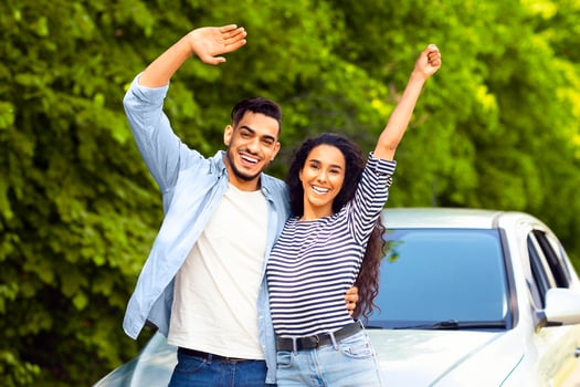 Emotional multiracial couple standing by nice auto at countryside, embracing and waving at camera, copy space. Happy middle-eastern man and woman having car trip together, renting automobile photo
