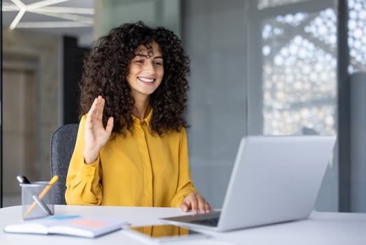Hispanic businesswoman participating in virtual meeting using laptop in professional office. Woman waves and smiles, conveying friendly communication. photo