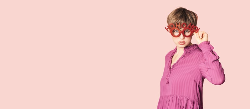 A cheerful young woman in Christmas glasses poses on a workspace layout highlighted on a simple pastel light pink background. photo
