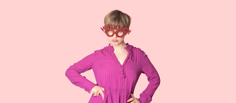 A cheerful young woman in Christmas glasses poses on a workspace layout highlighted on a simple pastel light pink background. photo