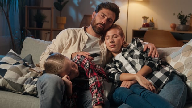 Tired sleepy exhausted fatigued Caucasian family mother woman man father with child kid girl daughter together sleeping napping relaxing on couch at home in evening dark night living room cozy sofa photo