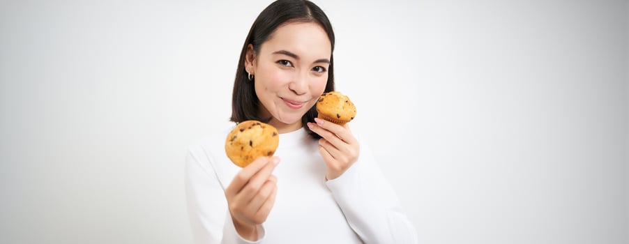 Image of korean woman enjoys eating baked pastry, showing two tasty cupcakes near face and smiles, white background photo