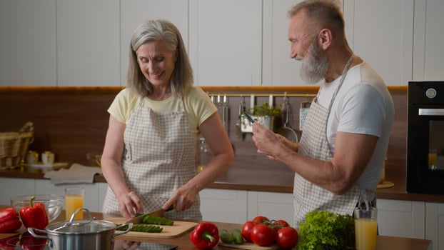 Retired old couple happy Caucasian family cooking in kitchen woman chef cutting cucumber chopping fresh healthy vegetables senior elderly man make photo on mobile phone cook on camera recording
