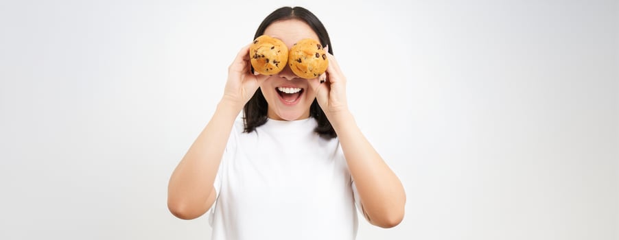 Close up portrait of funny asian girl, holds cupcakes near face, makes happy faces, white studio background photo