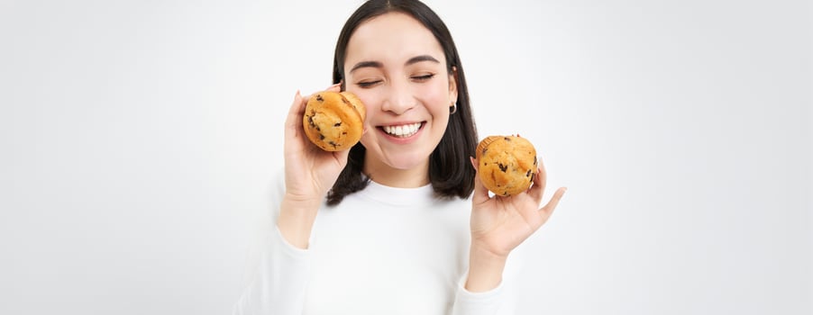 Close up portrait of asian woman, shows two cupcakes near face and smiles, likes pastry, enjoys eating bakery, white background photo