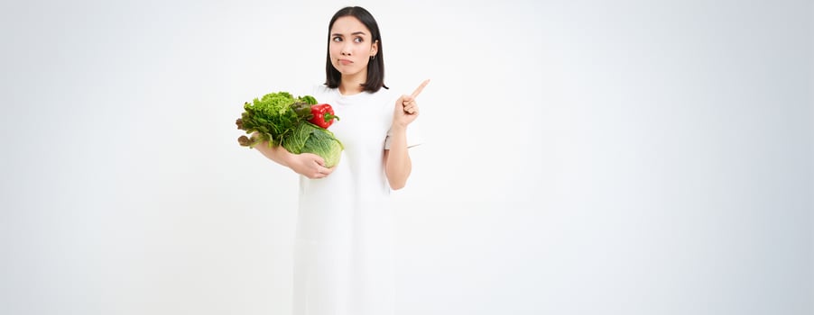 Portrait of pensive asian woman, holding vegetables, pointing right with thinking face, white background photo