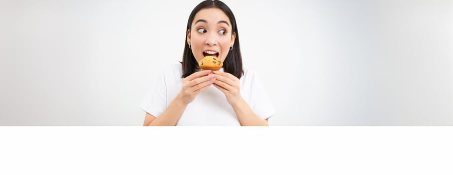 Close up of happy korean woman eating cupcake, delicious dessert, smiling from pleasure, white background photo