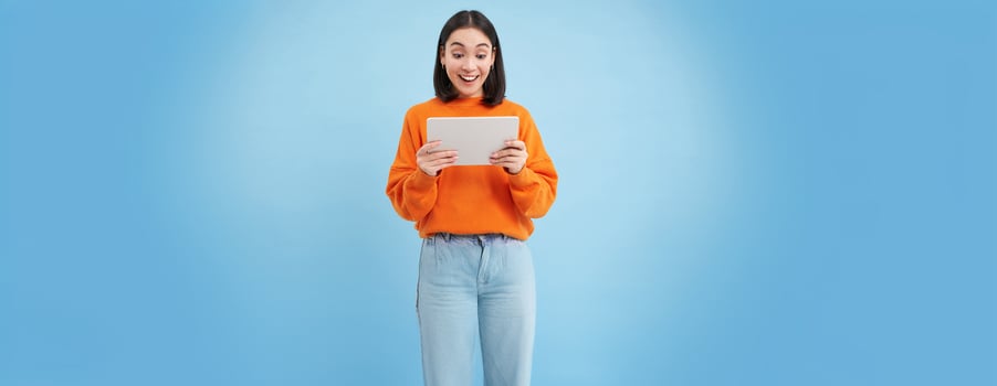 Smiling beautiful asian woman reading on digital tablet, watching videos on gadget or playing games, standing over blue background photo
