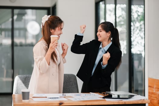 Two young businesswomen in a modern office celebrate success with coffee and documents, showcasing teamwork and achievement. photo