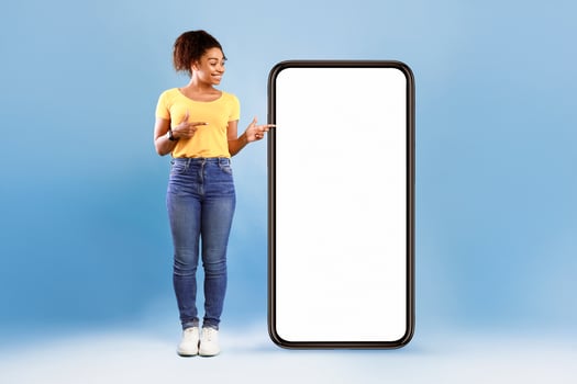 Cheerful African American woman standing next to huge smartphone, pointing at empty screen, advertising new mobile application or website. Blue studio background, cellphone display mockup photo