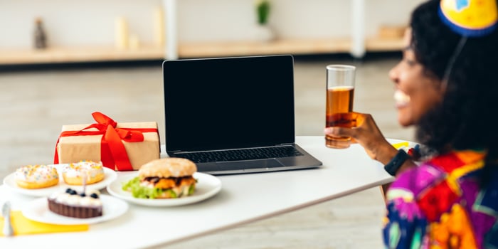 Back-View Of African Female In Festive Hat At Laptop With Blank Screen Celebrating Birthday Or New Year Holiday Calling Holding Glass And Having Dinner At Home. Selective Focus, Mockup photo