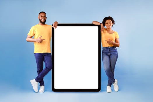 Hot offer, online sale. Cool African American young couple pointing at tablet pc with mockup space over blue studio background. Guy and lady advertising new awesome app or website photo