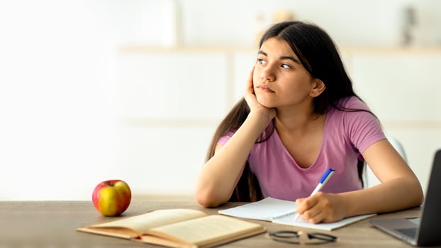 Thoughtful Indian teen girl studying online from home, taking notes during web lesson, feeling bored at home. Pensive adolescent participating in dull lecture, learning remotely during covid lockdown photo
