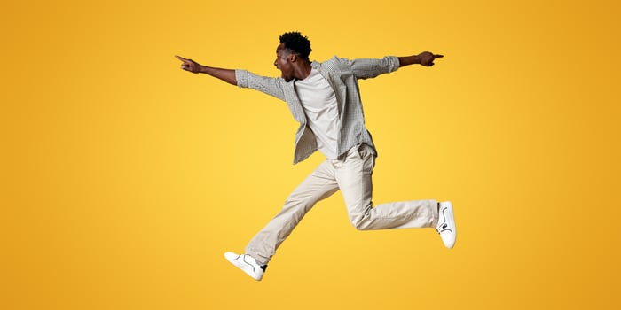 Crazy Sales. Excited black guy jumping up in air and pointing aside at copy space on beige background, emotional young african man demonstrating free place for design or advertisement, panorama photo
