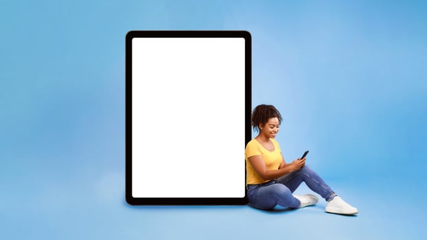 Full length of young African American lady using cellphone, sitting next to giant tablet pc with empty white screen, blue background. Mockup for app or website, space for advertisement design photo