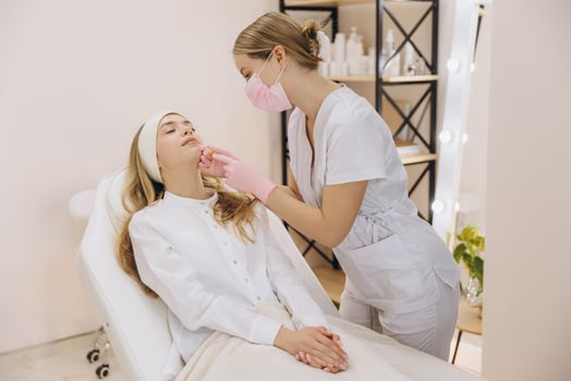 Beautician wearing pink surgical mask and gloves injecting botox in lips of young woman lying on white couch in beauty salon photo