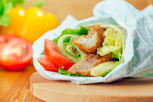 Fresh wrap filled with grilled chicken, lettuce, tomatoes, and cucumbers on a wooden surface with colorful vegetables in background photo