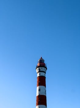 A striking red-and-white Osinovets Lighthouse stands tall against a vibrant blue sky, illuminated by sunlight. The historic maritime beacon, located on the shores of Lake Ladoga, is a significant photo