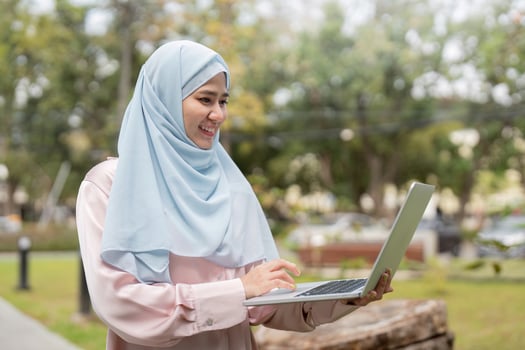Confident Muslim woman smiling while typing on a laptop in a park, showcasing professional engagement. photo