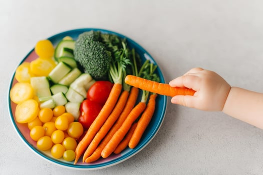 Toddler nutritious for child healthy concept. Colorful vegetables on a plate with a child's hand reaching for a carrot. photo