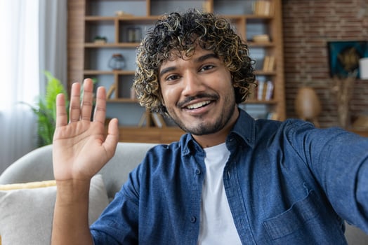 A man looks into the phone camera, happily waves his hand, takes a selfie photo and talks to colleagues and friends remotely using a smartphone app for chats