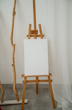 Wooden easel with blank canvas against white wall. Nearby is wooden decorative element. Vertical photo. Easel is on gray floor photo