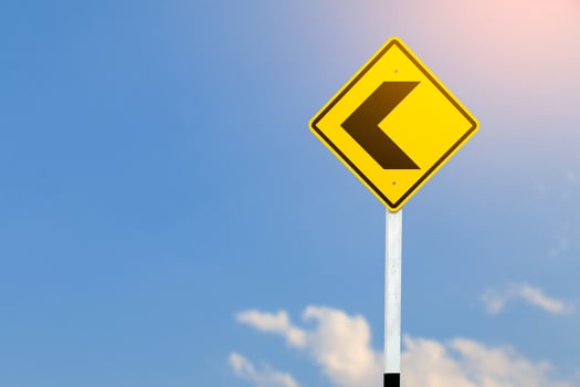 Black left direction arrow is displayed on a bright yellow traffic sign on blurred sky background. photo
