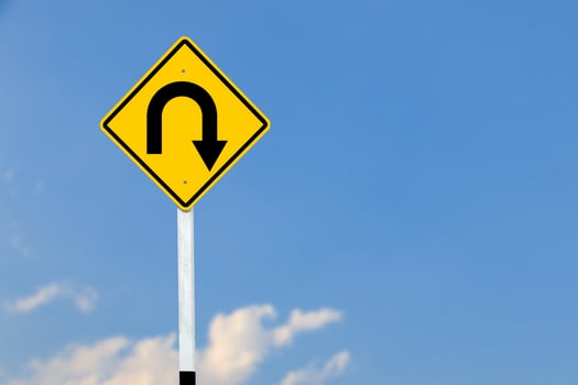 A black U turn symbol is prominently displayed on a bright yellow sign on blurred sky background. photo
