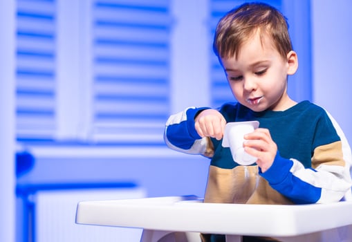 Child enjoys yogurt in a bright room. A young boy sits at a high chair, tasting yogurt with a spoon while basking in natural light. photo