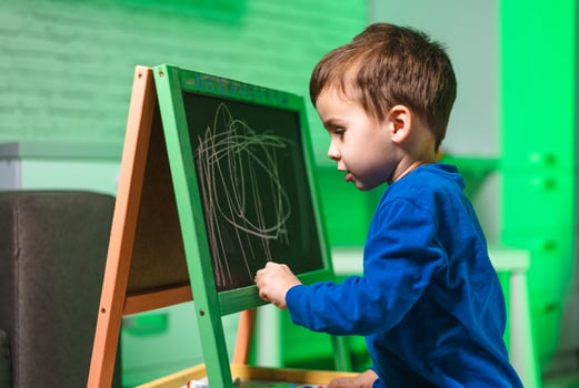Child creatively draws on board. A young boy in a blue shirt sketches on a green chalkboard with focused enthusiasm in a cozy room setting. photo
