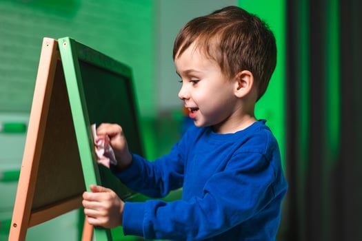Kid joyfully erasing board. A young boy in a blue shirt joyfully wipes a chalkboard in a bright, creative room. photo