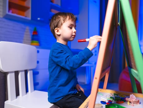 Young boy painting creatively at home. A young boy is focused on painting at his easel in a bright room filled with colorful decorations and toys. photo