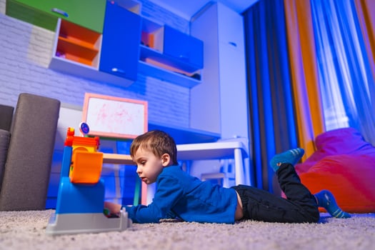 Child plays indoors. A young boy lies on a soft carpet, focused on a colorful toy. The room is brightly lit with blue and orange hues. photo