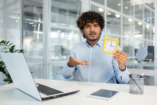 Young man at his office desk holding a yellow square clock, showing his concern about running late or managing time effectively. photo