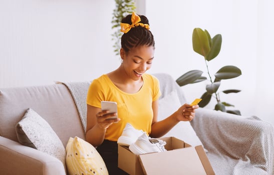 Online Shopping And Payment. Portrait of excited african american female buyer holding credit card and using mobile phone, sitting on couch and looking in cardboard box container with white sneakers photo